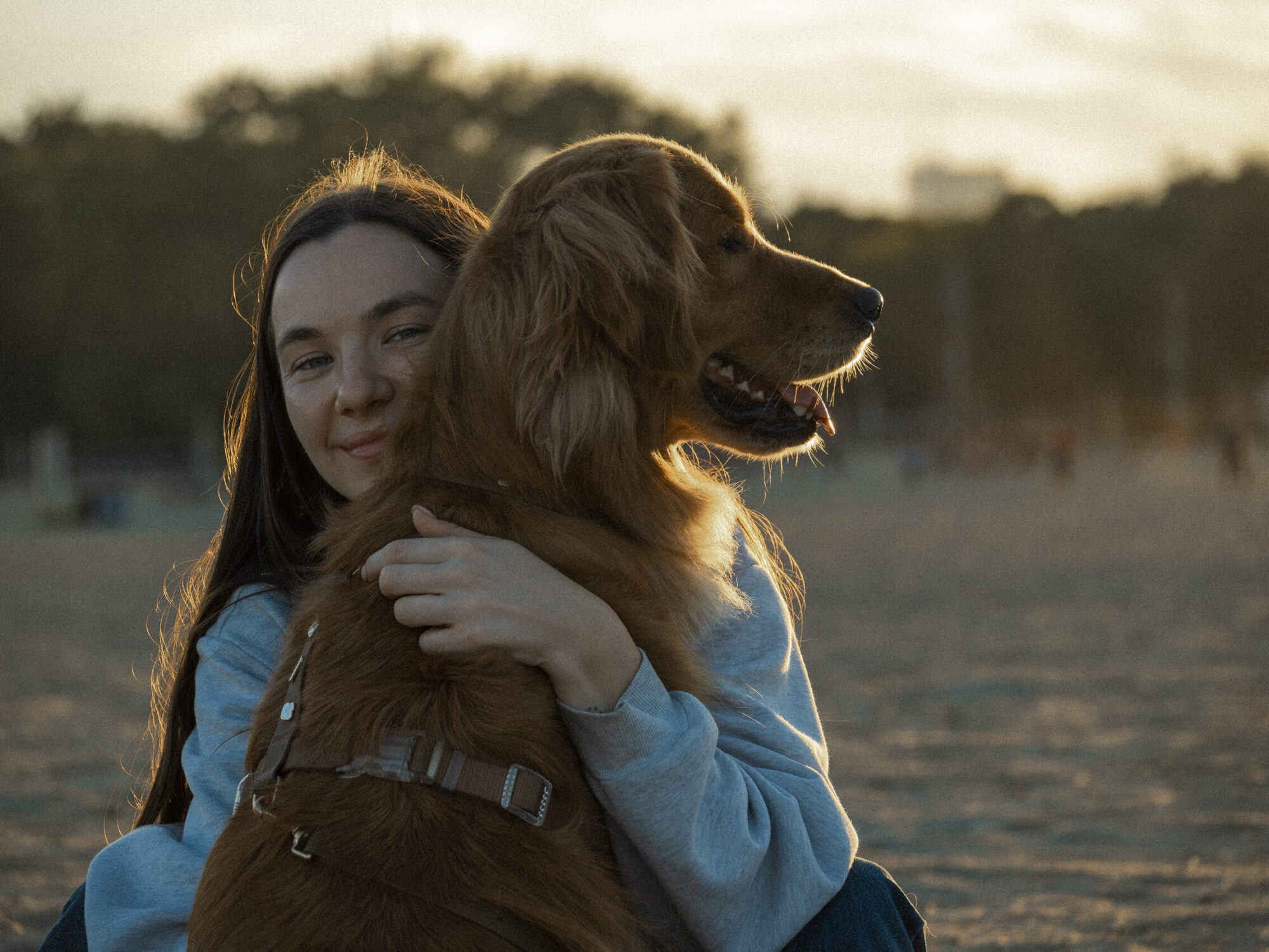 ferm-emelie_DSC_1185 Woman sitting on the beach at sunset, hugging her dog while looking in the camera.