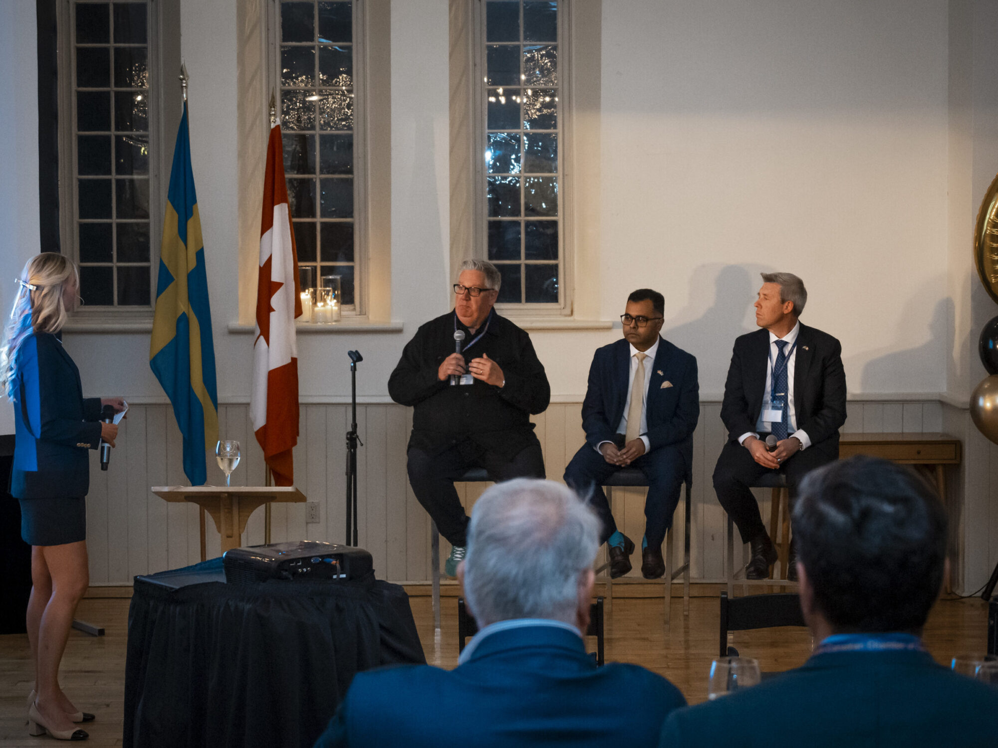 ferm-emelie_DSC2245_sccc-60th Wide view of a panel discussion with three men seated and a woman standing as the host.