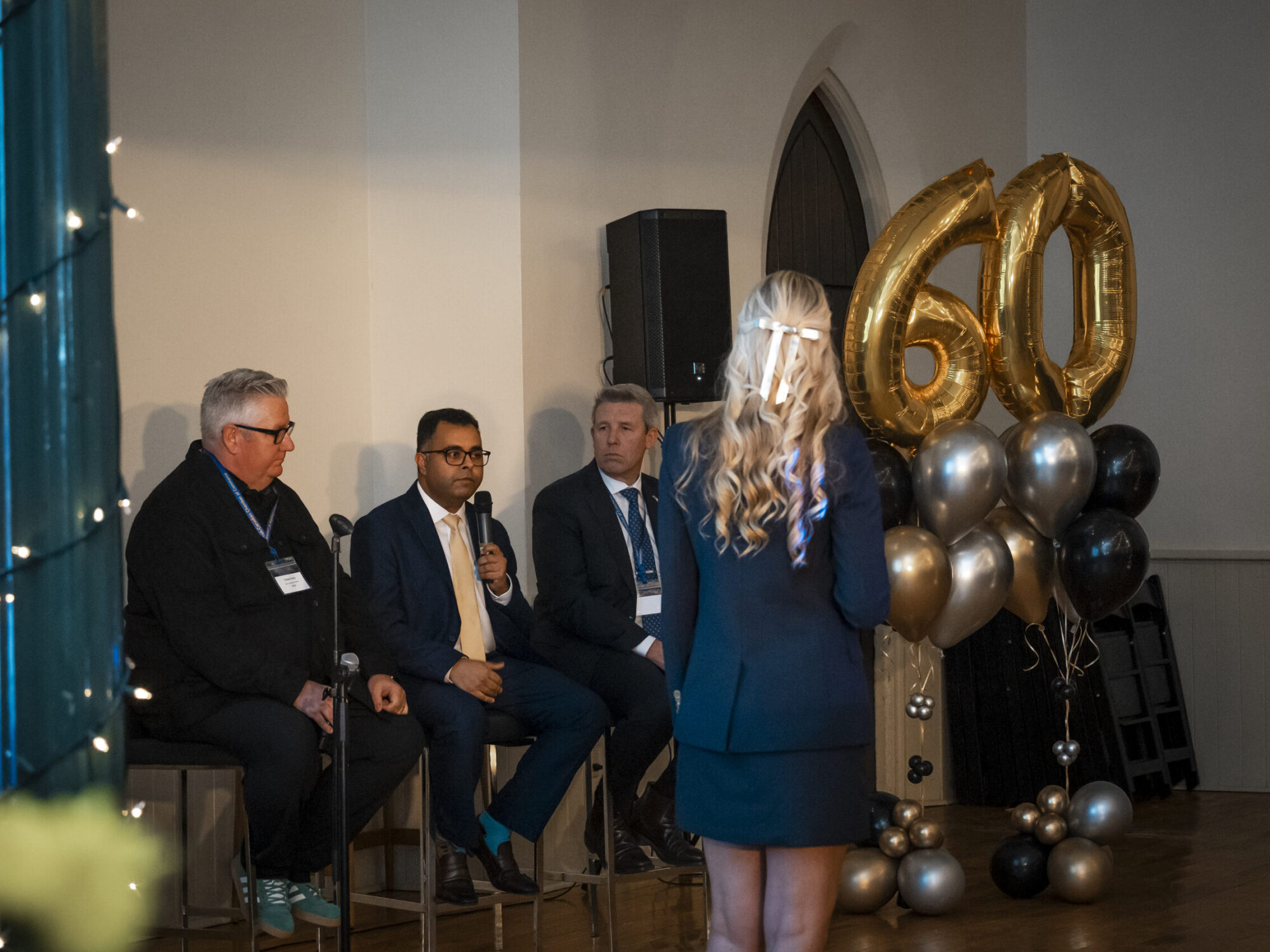 ferm-emelie_DSC2238_sccc-60th Wide view of a panel discussion with three men seated and a woman standing as the host.